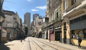 Imagem de uma rua do Centro do Rio de Janeiro durante o dia. Alguns prédios com 2 ou 3 pavimentos do lado direito e esquerdo e prédios maiores no fundo.